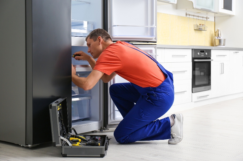 Male Technician With Screwdriver Repairing Refrigerator In Kitchen