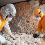 Two workers in protective suits and masks handling debris indoors, likely during a hazardous material cleanup or asbestos removal in a damaged building.